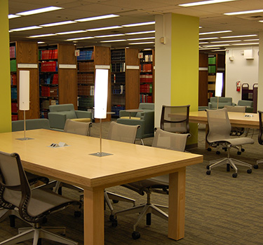 Large open area with lights on wooden tables surrounded by six wheeled chairs and cushioned chairs.