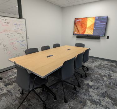 Room with a table, chairs, whiteboard and large monitor on the wall.