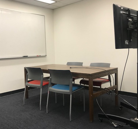 Study room with a wood table with four chairs around it, large television screen on a stand, and a whiteboard on the wall